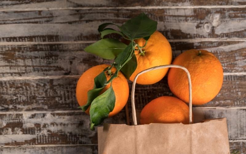A brown paper bag with a handle laying on a wooden background with oranges s