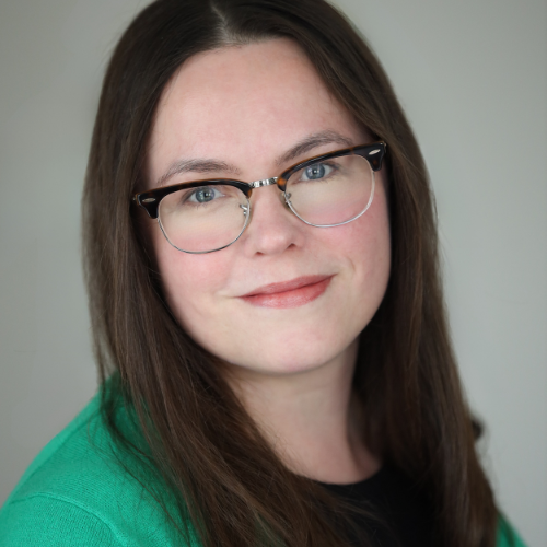 A white woman with dark hair and glasses is wearing a dark shirt and a green cardigan.