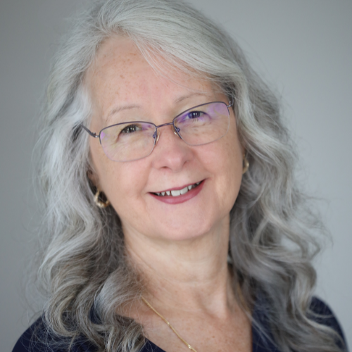 A white woman with white hair and glasses is smiling at the camera. She is wearing a dark shirt and a gold necklace.