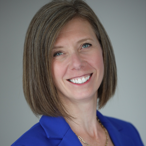 A white woman with brown hair is smiling. She is wearing a blue blazer.