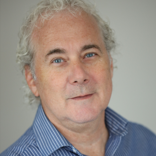 A white man with white curly hair is looking at the camera. He is wearing a blue dress shirt.