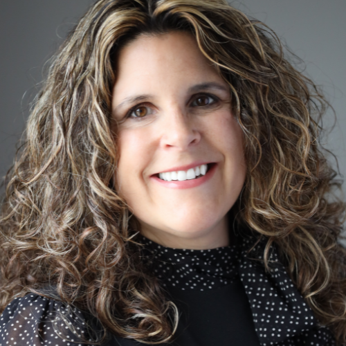 A white woman with brown curly hair is wearing a black shirt with polka dot sleeves and scarf.