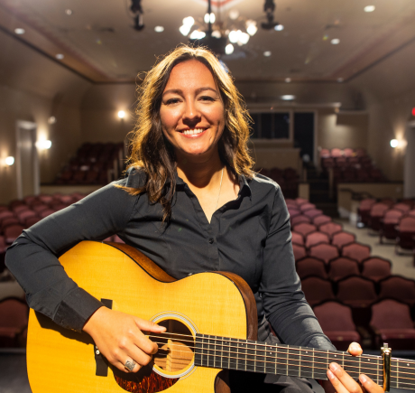 A white woman with dark hair is sitting on a stage and holding a guitar.