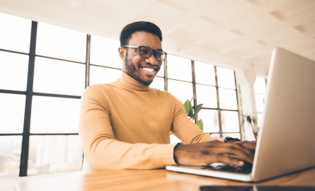 A black man wearing glasses and a turtleneck is working on a laptop.