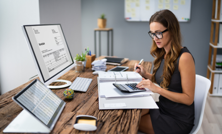 A woman sitting at a desk with a computer in front of her is working on a calculator.