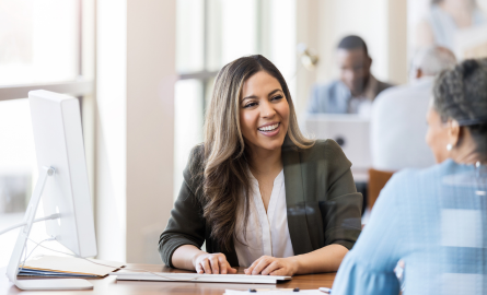 A woman working on a computer is smiling at a woman across her desk.