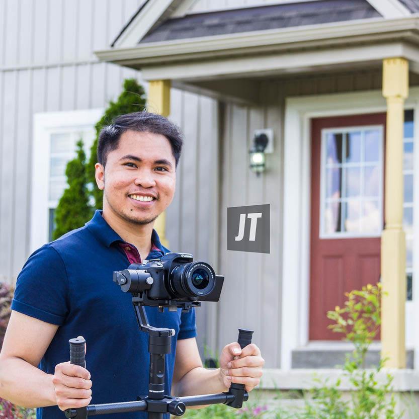 Young dark haired man is looking at the camera. He is wearing a blue shirt and holding a camera on a tripod.