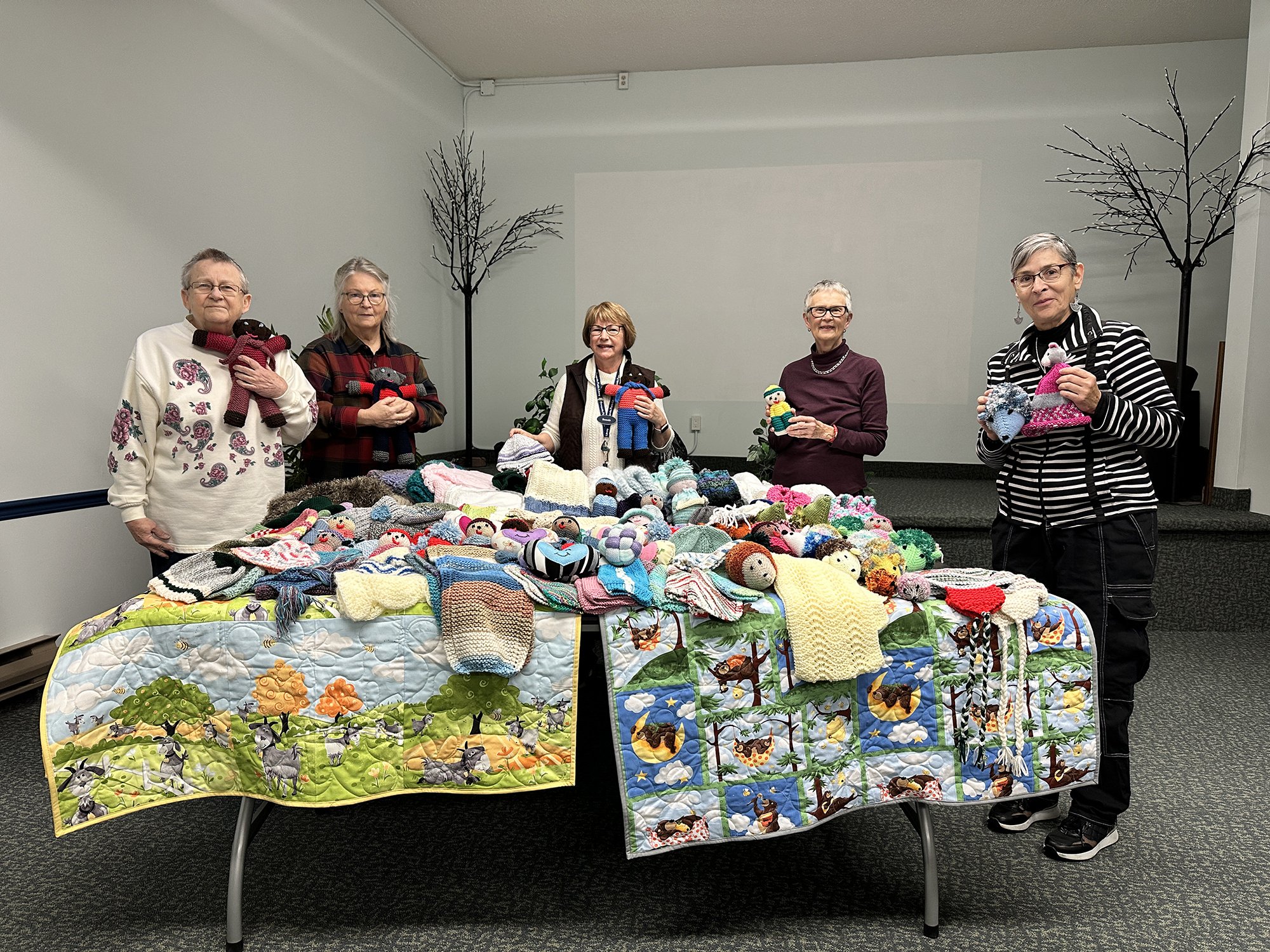 Members of the Orange thREADs fabric artist group stand behind a table of knitted, crocheted, and quilted donations