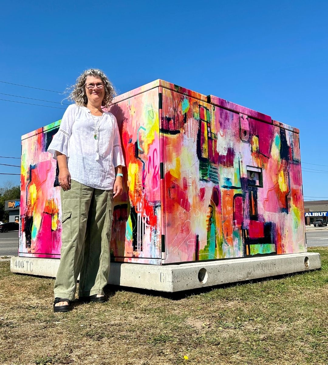Spirit of Community by Deb Menken A woman standing beside of Hydro transformer covered in abstract artwork