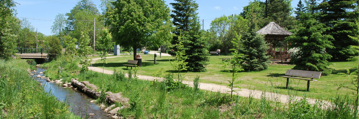 Kay Cee Garden park. Image view is a wide span of the park showing a river, benches, mixed trees and a gazebo.