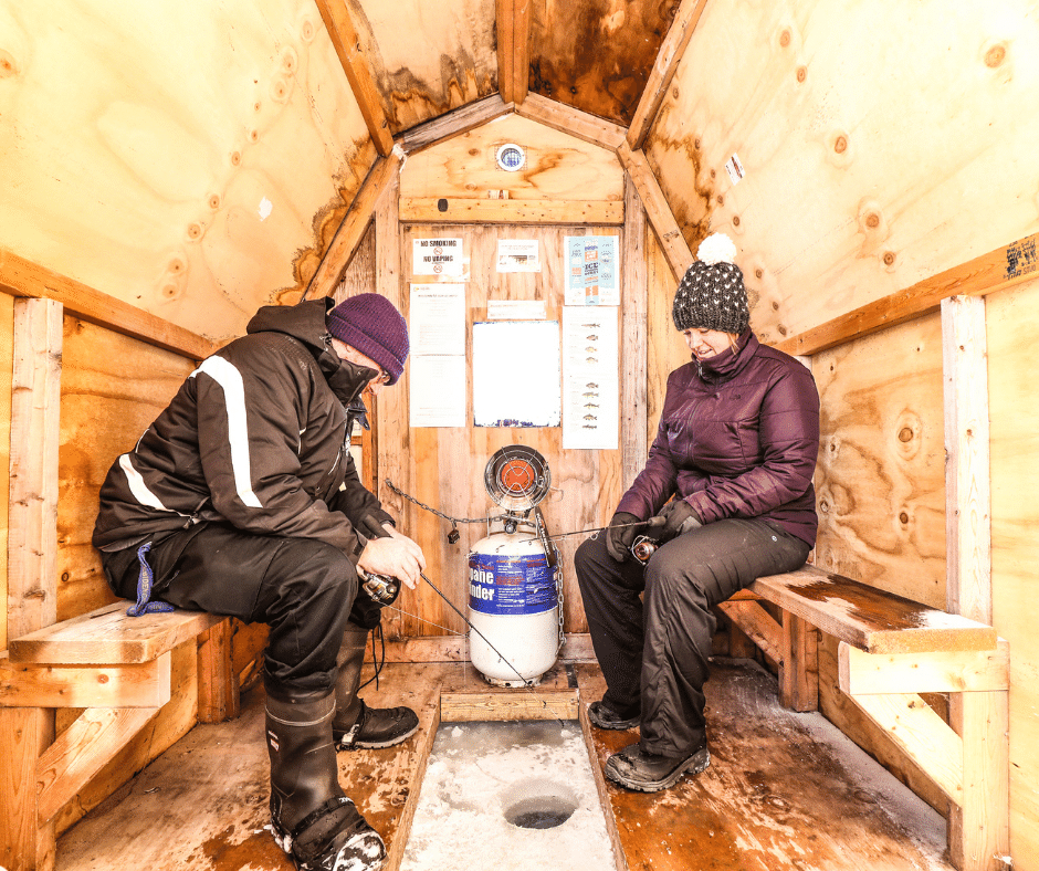 Man and a women sitting inside of a wooden ice fishing hut. Both are wearing snowsuits and folding fishing rods.