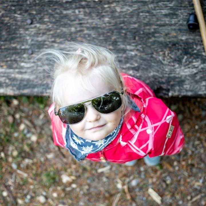 Young blonde girl wearing sunglasses and a pink snowsuit looking up at the camera
