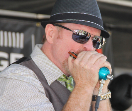 A white man is wearing a black fedora and sunglasses. He is playing a harmonica.