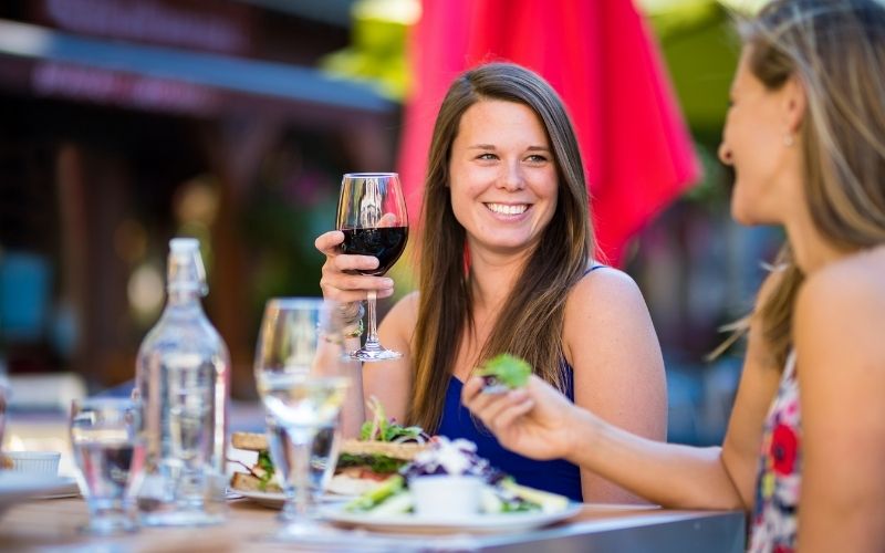 A dark haired white woman in a blue tank top is holding a glass of red wine. She is sitting at a patio table in the summer time.