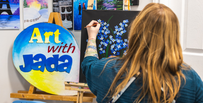 A woman has her back to us while she paints blue flowers on a black canvas
