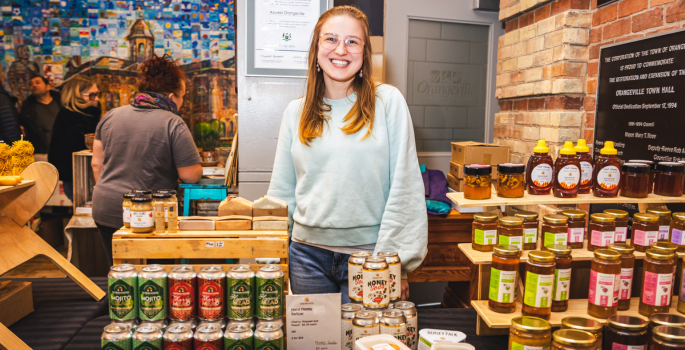 A woman stands behind a counter at a market, selling honey items.