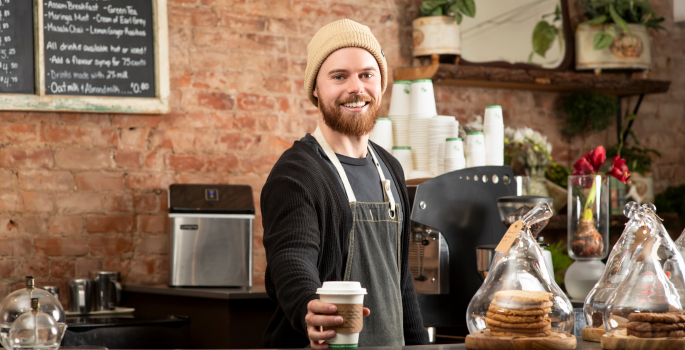 A man stands behind the counter at a cafe. He is handing a customer a coffee.