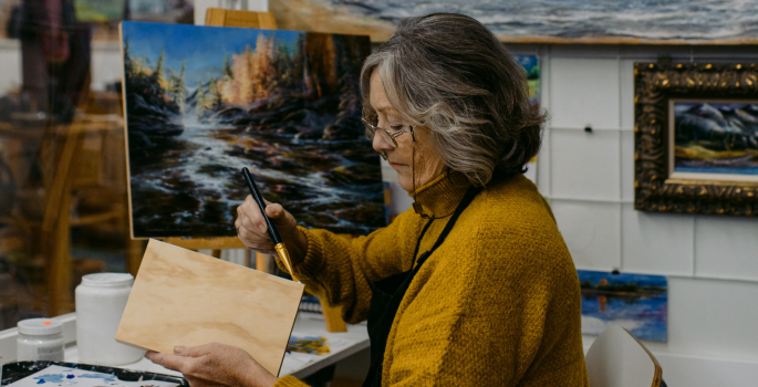 A woman sits and paints while surrounded with paintings.