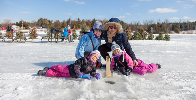 Mother, Father and two children say on a, ice covered lake with a fishing hole in front of them.