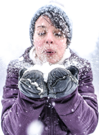 A woman dressed in a purple winter jack is blowing a handful of snow in her mittens.